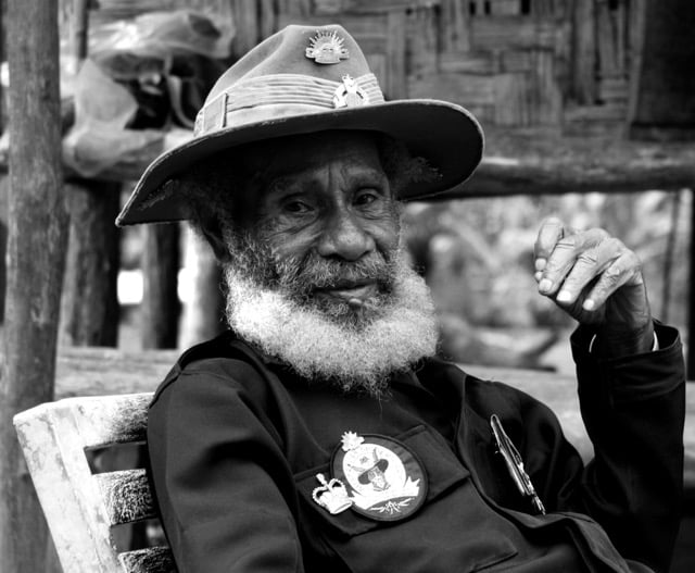 A black and white portrait of an elderly man with a white beard wearing a traditional military slouch hat and a jacket adorned with badges, seated outdoors.