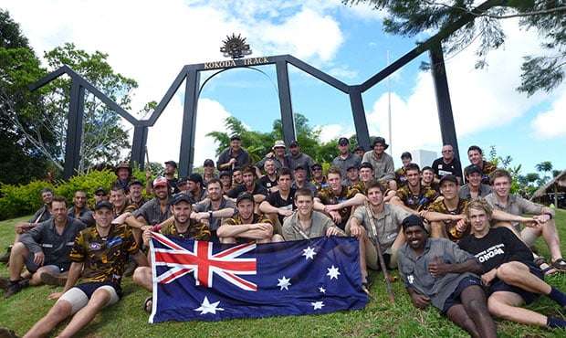 A large group of men in athletic and hiking gear posing for a photo in front of the black arches of the Kokoda Track memorial in Papua New Guinea. Several men in the front row are holding a large Australian flag.