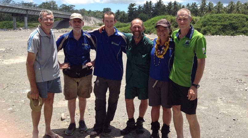 Six adults—five men and one woman—standing in a line with their arms around each other. They are dressed in casual hiking or outdoor gear. In the background, there is a bridge, green foliage, and palm trees under a bright, sunny sky.