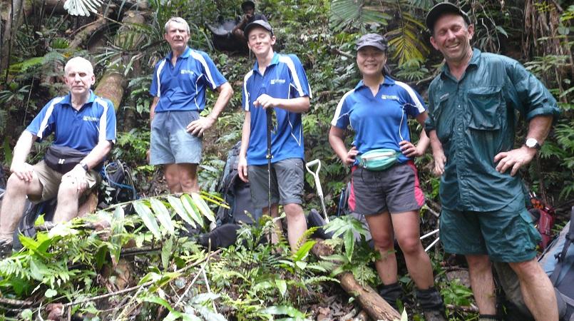 A group of five hikers posing on a lush, tropical trail. Three men and one woman wear matching blue athletic shirts, while a fifth man on the right wears a dark green button-down. They are surrounded by dense ferns and trees.