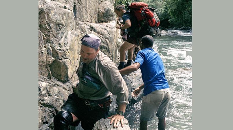 A group of trekkers and a local guide carefully scramble over steep rocks alongside a fast-flowing jungle river.
