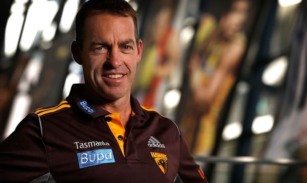 A medium close-up of a smiling man wearing a dark brown and orange polo shirt with "Tasmania," "Bupa," "Adidas," and "Hawks" branding.