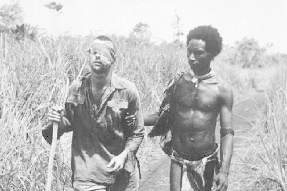 A black-and-white historical photograph depicts a local indigenous man assisting a wounded, blindfolded soldier through tall grass.