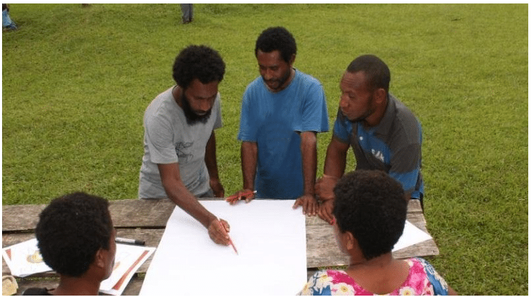 A group of local men and women gather around a wooden table outdoors, focused on a large white map or planning document.
