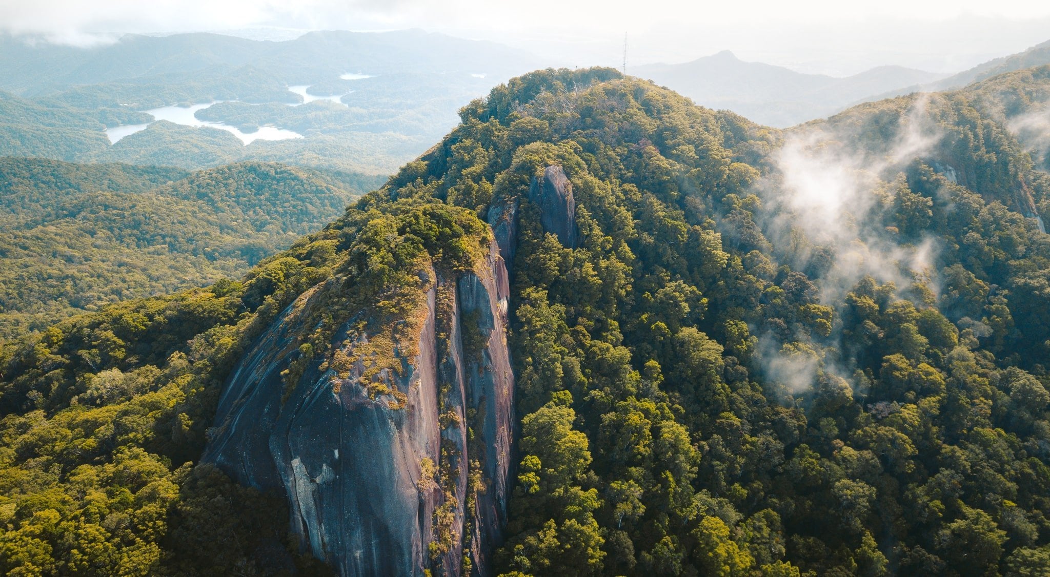 An aerial view of a massive granite rock face rising out of a dense green forest, with mist clinging to the trees and a winding river in the distance.