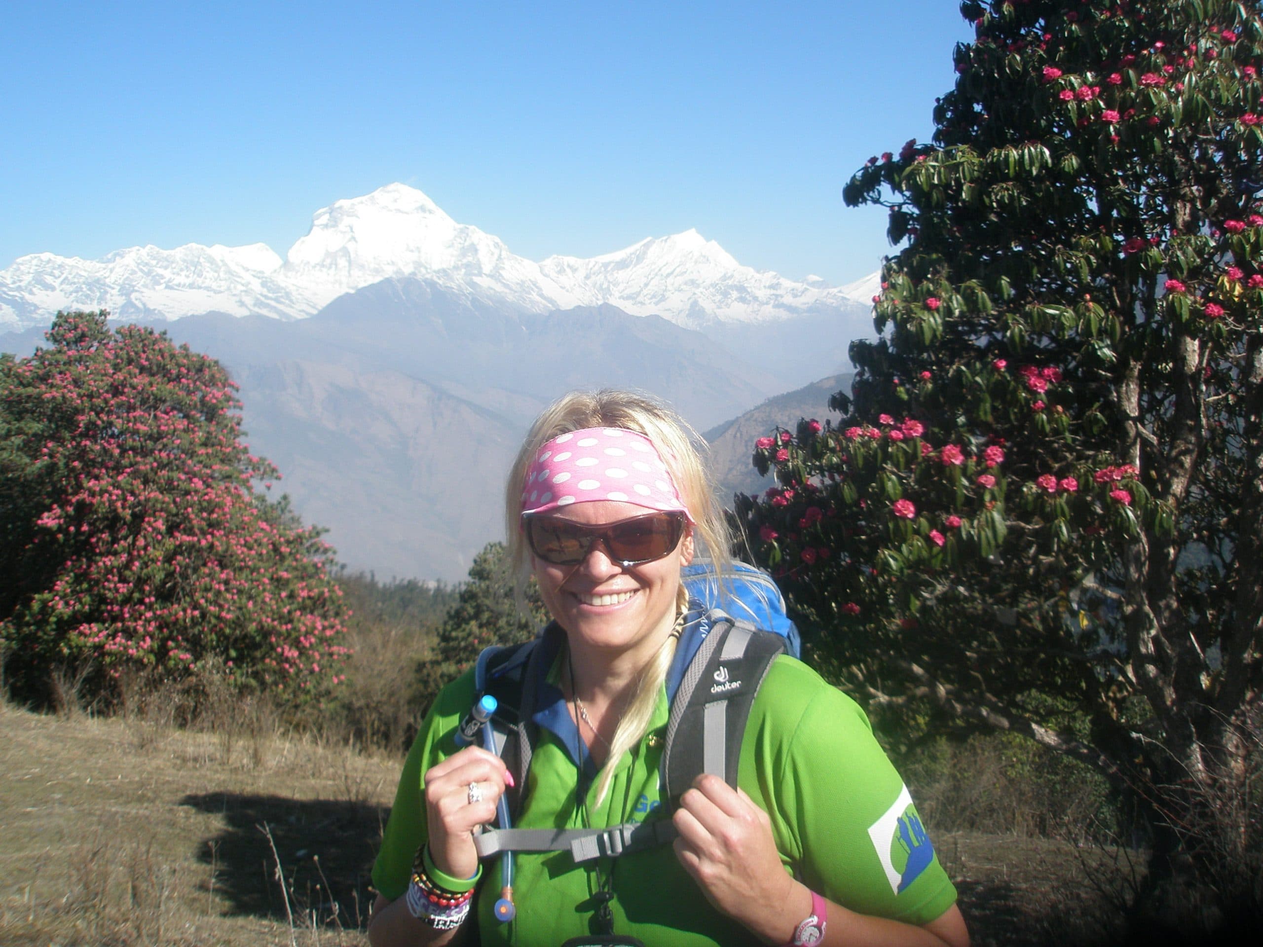 A woman wearing a green trekking shirt, sunglasses, and a pink polka-dot headband smiles at the camera. She is surrounded by lush trees with vibrant pink flowers. In the background, sharp, white peaks rise above a blue mountain haze under a clear sky.