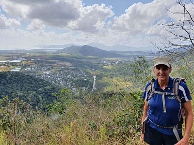 A smiling female hiker in a blue shirt standing on a paved road curving through a lush forest.