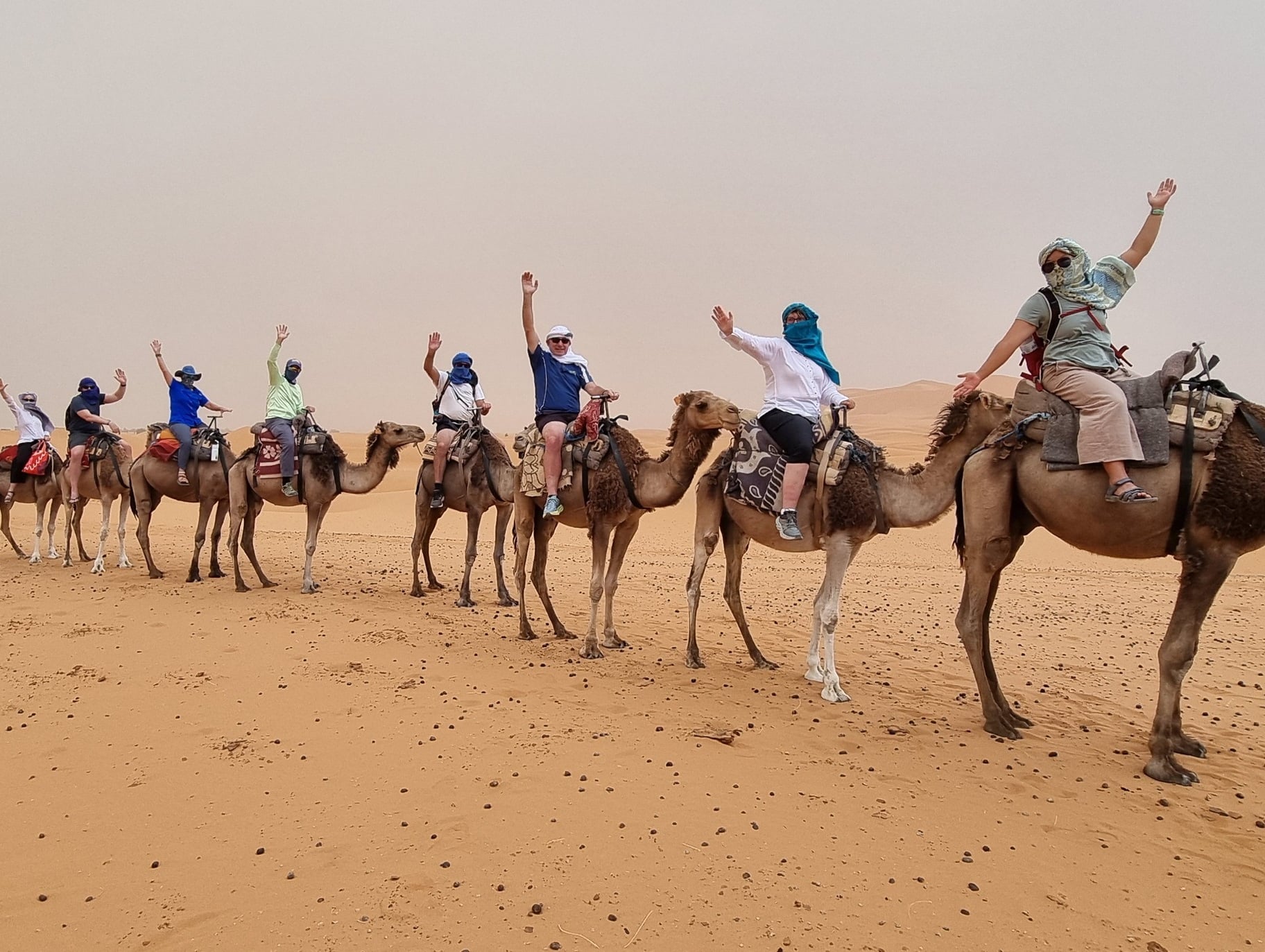 Group of eight people riding camels in desert; each person raises one arm, posing or waving; riders wear light protective clothing with headscarves and sunglasses; camels equipped with saddles and riding gear; vast sandy landscape with distant dunes under hazy sky.