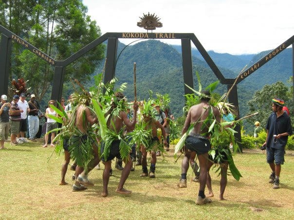 A group of performers wearing traditional attire made from leaves and natural materials dance in front of the Kokoda Trail archway. The archway bears inscriptions honoring "Papuan Carriers" and the "Koiari People." Spectators watch from the sidelines, with lush green mountains and dense trees filling the scenic background. Would you like a version optimized for SEO or accessibility standards? I can tailor it for image search discoverability or screen reader clarity.