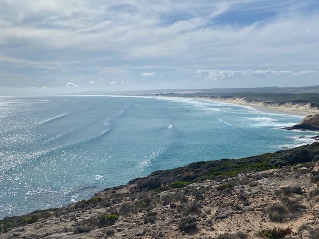 Expansive coastal landscape with sandy beach curving into the distance, gentle waves meeting the shore; rocky terrain with sparse vegetation in the foreground, rolling hills in the background under a partly cloudy sky.