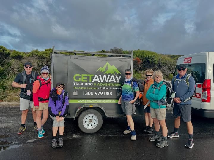 Group of seven hikers in trekking gear standing in front of a trailer and a van; trailer sign reads “GETAWAY TREKKING & ADVENTURES – The Aussie Camino, Portland to Penola” with contact details; van beside it displays the AVIS rental logo.