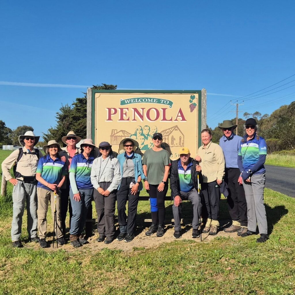 Group of twelve people in hiking gear standing in front of a large roadside sign reading “WELCOME TO PENOLA”; illustrated sign includes people, buildings, and a bunch of grapes symbolizing vineyards; rural setting with grass, trees, and power lines under a clear blue sky.