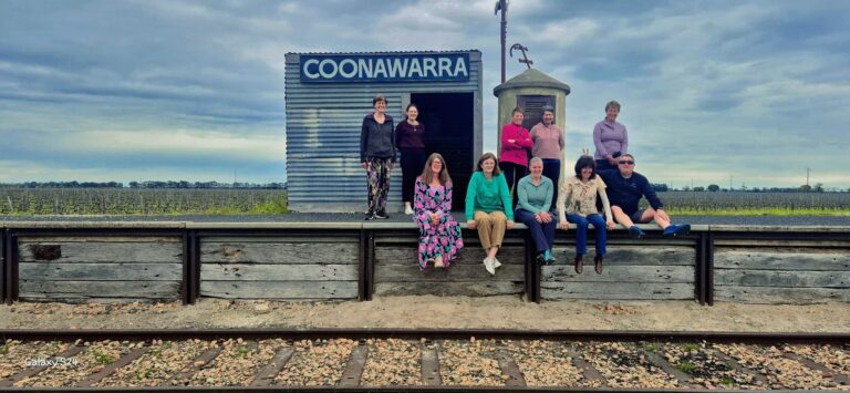 Group of ten people posing on a rustic train station platform labeled “COONAWARRA”; corrugated metal building and cylindrical structure beside the platform; wooden planks elevate the platform next to railway tracks; background shows expansive vineyard under a cloudy sky; group casually dressed, some standing and others sitting on the edge of the platform.