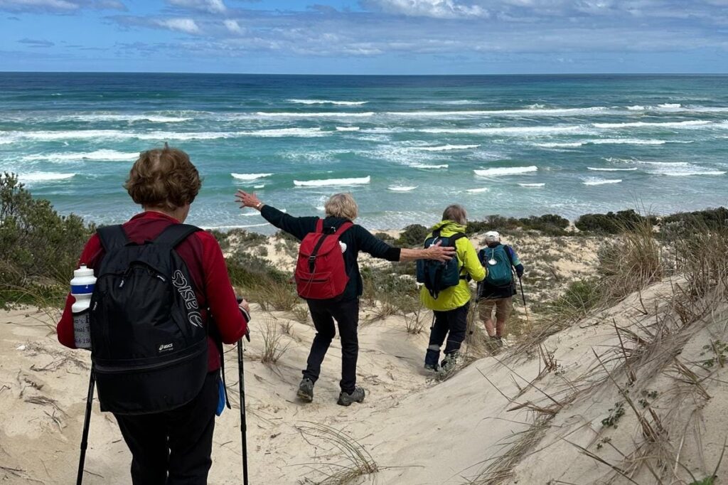 Four friends hiking through sand dunes toward a vast, sparkling blue ocean. The leader of the group has their arms wide open, soaking in the coastal view and the sea breeze.