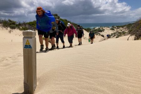 A line of hikers with backpacks climbs a tall, rippled sand dune toward a trail marker under a cloudy sky.