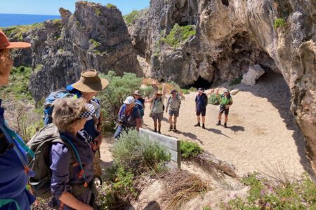 A group of hikers waving from a sandy area in front of a large, rugged sea cave along a rocky coastline.