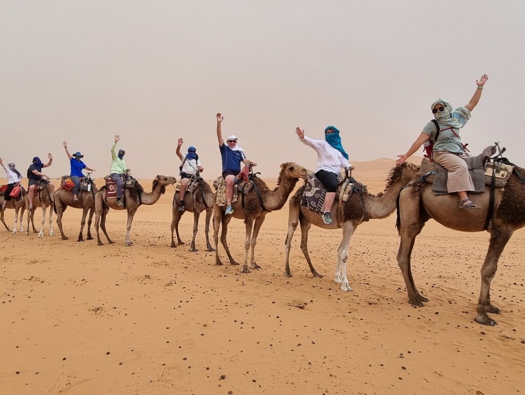 Group of seven people riding camels in desert; each rider raises one arm in gesture of excitement or celebration; camels arranged in single line; riders wear head coverings and casual clothing suited for desert; expansive sand dunes stretch into background under hazy sky.