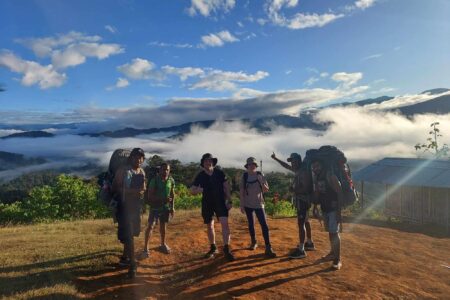 Group of six hikers standing on dirt clearing with backpacks; dressed in trekking gear; scenic mountainous landscape with lush green vegetation and dramatic mist settled in valleys behind; sky mostly clear with scattered clouds; small wooden structure with metal roof visible to right; scene conveys adventure, camaraderie, and natural beauty during hiking expedition.