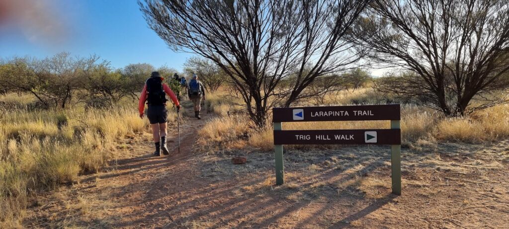 A group of hikers follows a dirt path into the scrubland, passing a brown wooden sign for the "LARAPINTA TRAIL" and "TRIG HILL WALK".