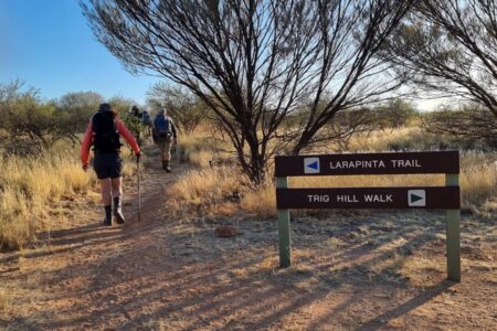 A group of hikers follows a dirt path into the scrubland, passing a brown wooden sign for the "LARAPINTA TRAIL" and "TRIG HILL WALK".
